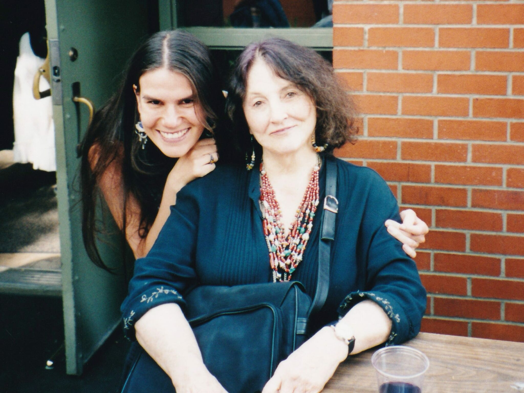 A woman (Alexandra Hidalgo) is standing and she leans forward to put her arms around a woman who (Bobbie Lousie Hawkins) is sitting at a table. They smile at the camera. They are outside with brick walls behind them. It's afternoon.