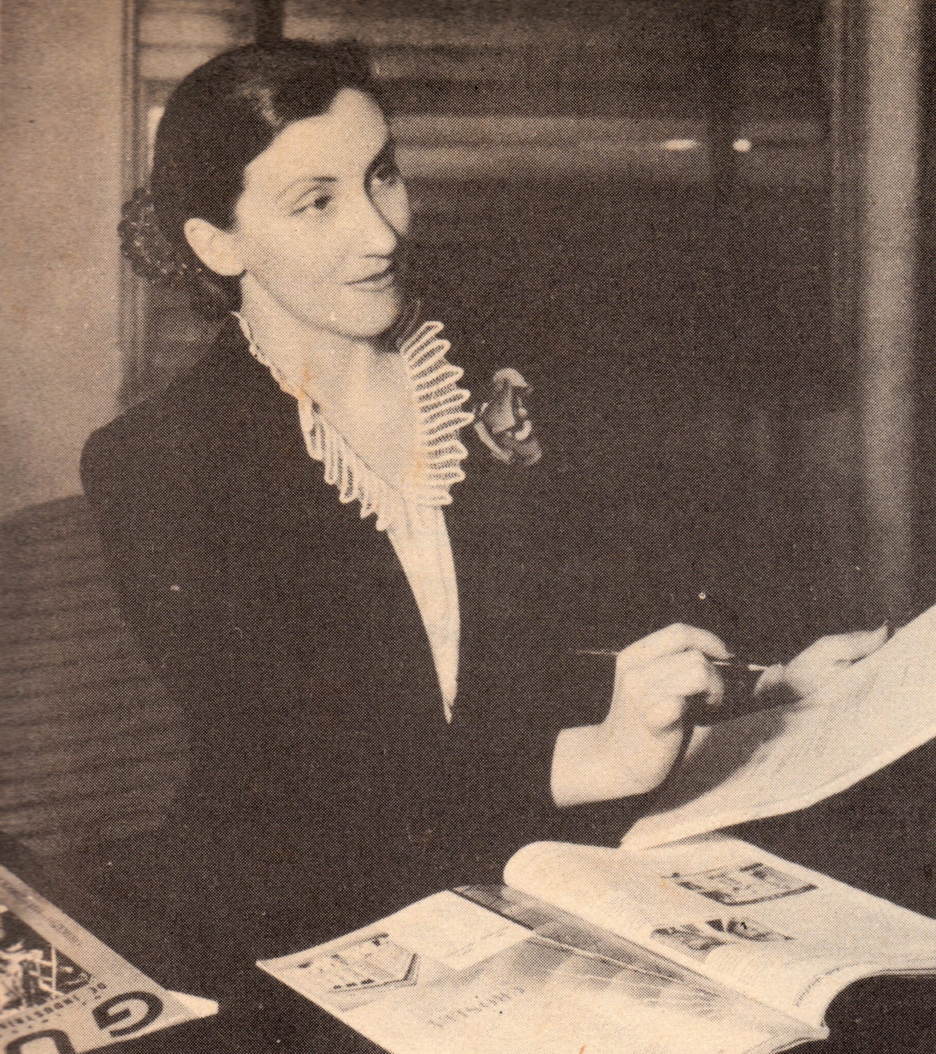 A woman (Olga Briceño) sits at a stately desk in this black and white image that is granulated from having been scanned from a paper brochure. She is gazing into space, pen and paper in her hand. The image is from the 1940s or so.
