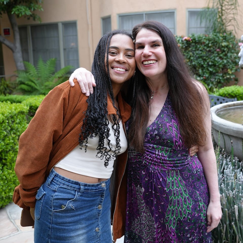 Two women pose for a picture to the left of a fountain. They are standing next to each other in an embrace, smiling with their faces pressed together. Behind them are bushes and foliage in front of a tan wall, they are outside, and it is daytime.