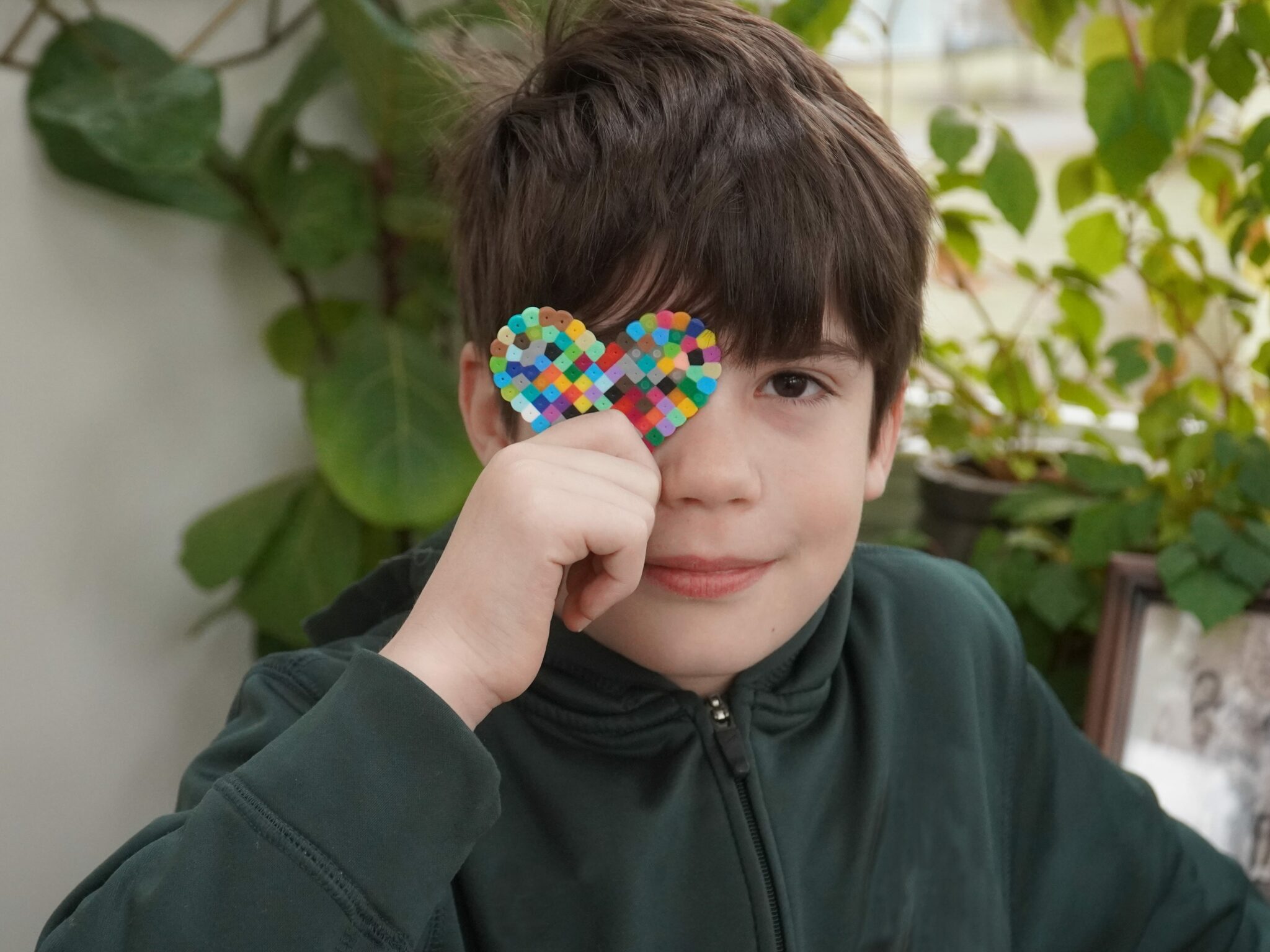 A boy smiles at the camera. Hi is holding a multicolored heart to his right eye, playfully hiding it. Behind him there are plants in soft focus. It is daytime.