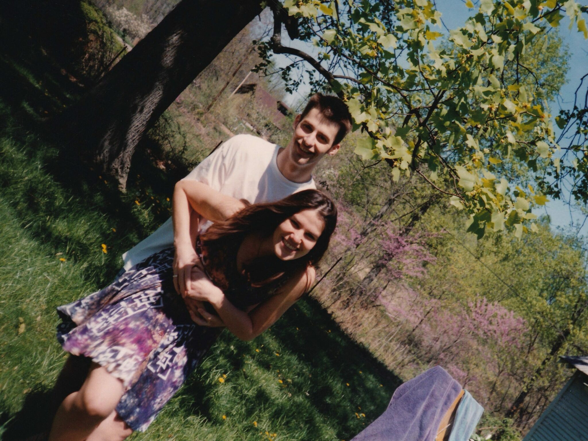 A man (Nathaniel Bowler) hugs a woman (Alexandra Hidalgo) from behind as they both smile at the camera. They are outside in a yard with a tree behind them. There are purple and white bushes and dandelions behind them. It’s daytime and the light is harsh.