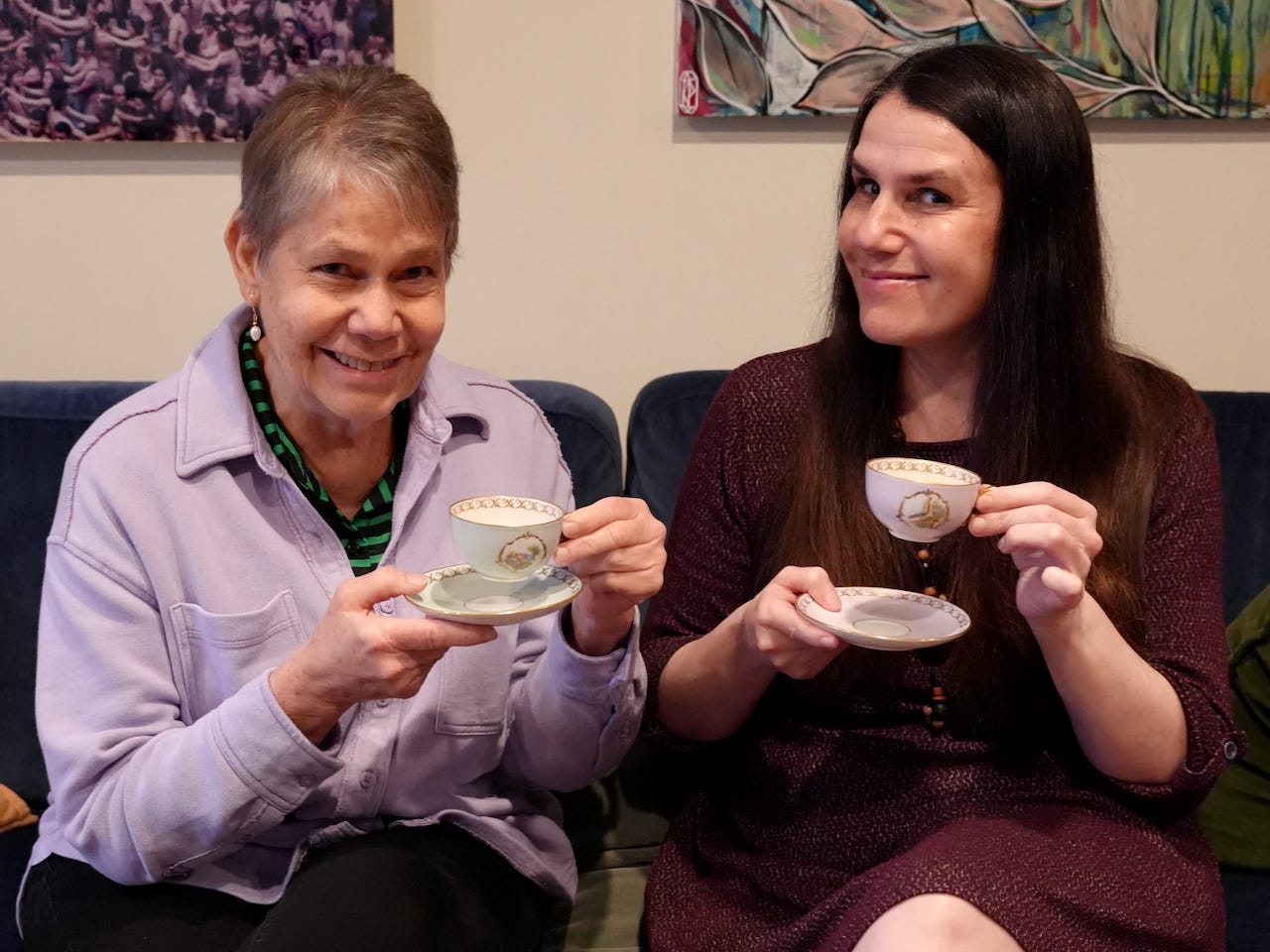 Two women, Antonieta Aagaard (left) and Alexandra Hidalgo (right), sit on a couch, smiling. They are holding tea cups. There are cream-colored walls behind them. It is daytime.