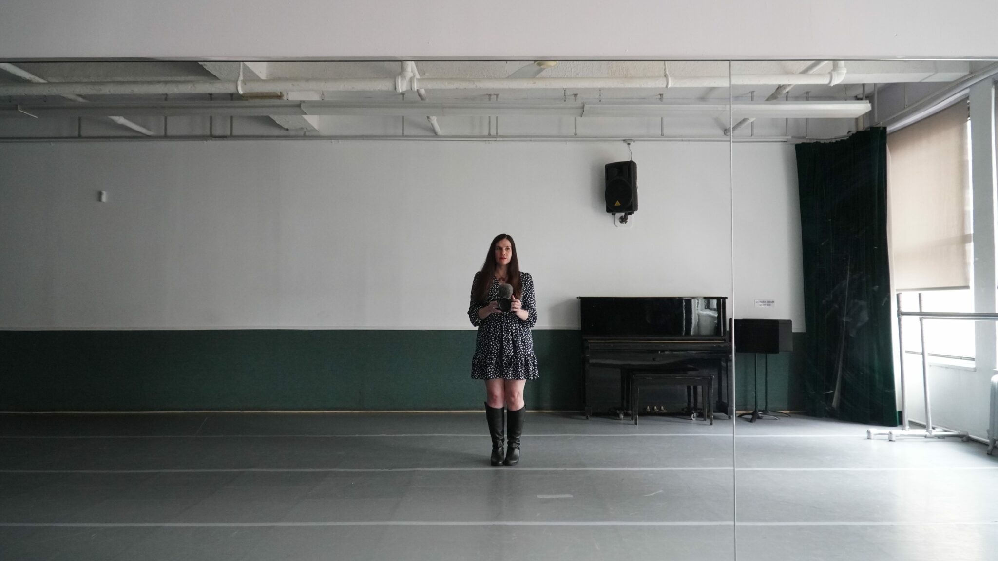 A woman stands in front of a mirror in a rehearsal room. She looks tense and stands to the side. The room is empty and there is a piano behind her.