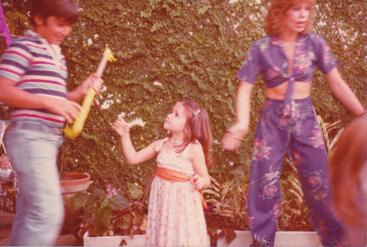 In a photo faded from time, a four year old girl looks at a much taller boy, her hand extended toward the piñata stick he is holding. Be her side is a woman in purple pants and a tiny matching top. She indeed looks sexy. It is daytime. They are outside and there’s a wall covered with green plants behind them.