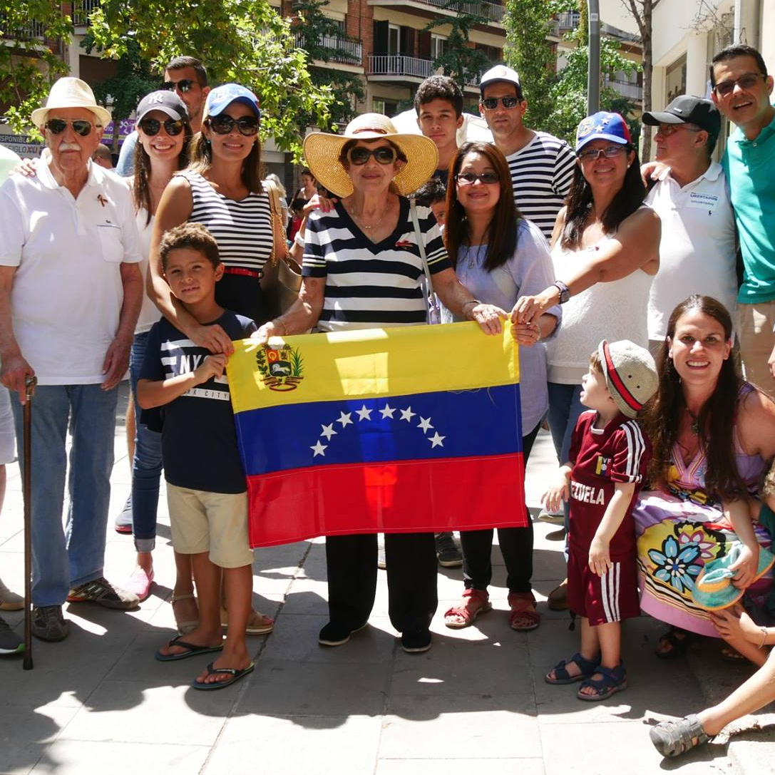 A group of Venezuelans stand together in Barcelona, Spain in 2017 during an anti-Maduro protest. A woman at the center is holding a Venezuelan flag. They smile at the camera.
