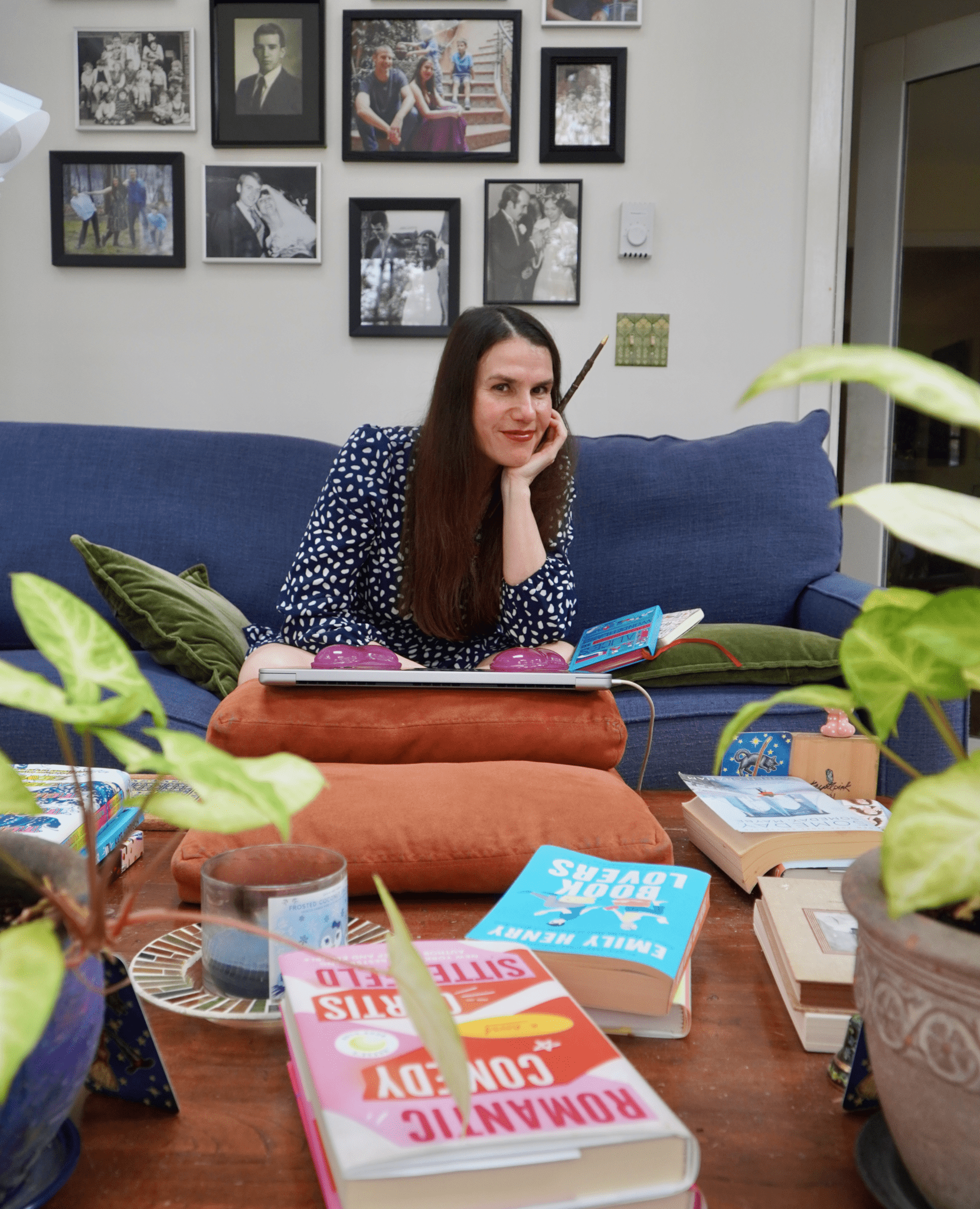 A woman is sitting in her living room while an array of books surround her. She holds a pen as she smiles into the camera.